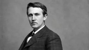 Thomas Edison trabaja en un laboratorio histórico rodeado de lámparas incandescentes, herramientas eléctricas, cables y aparatos experimentales.