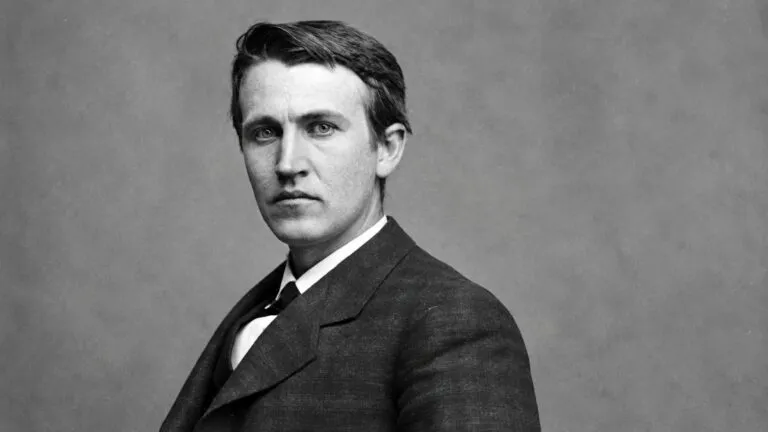 Thomas Edison trabaja en un laboratorio histórico rodeado de lámparas incandescentes, herramientas eléctricas, cables y aparatos experimentales.